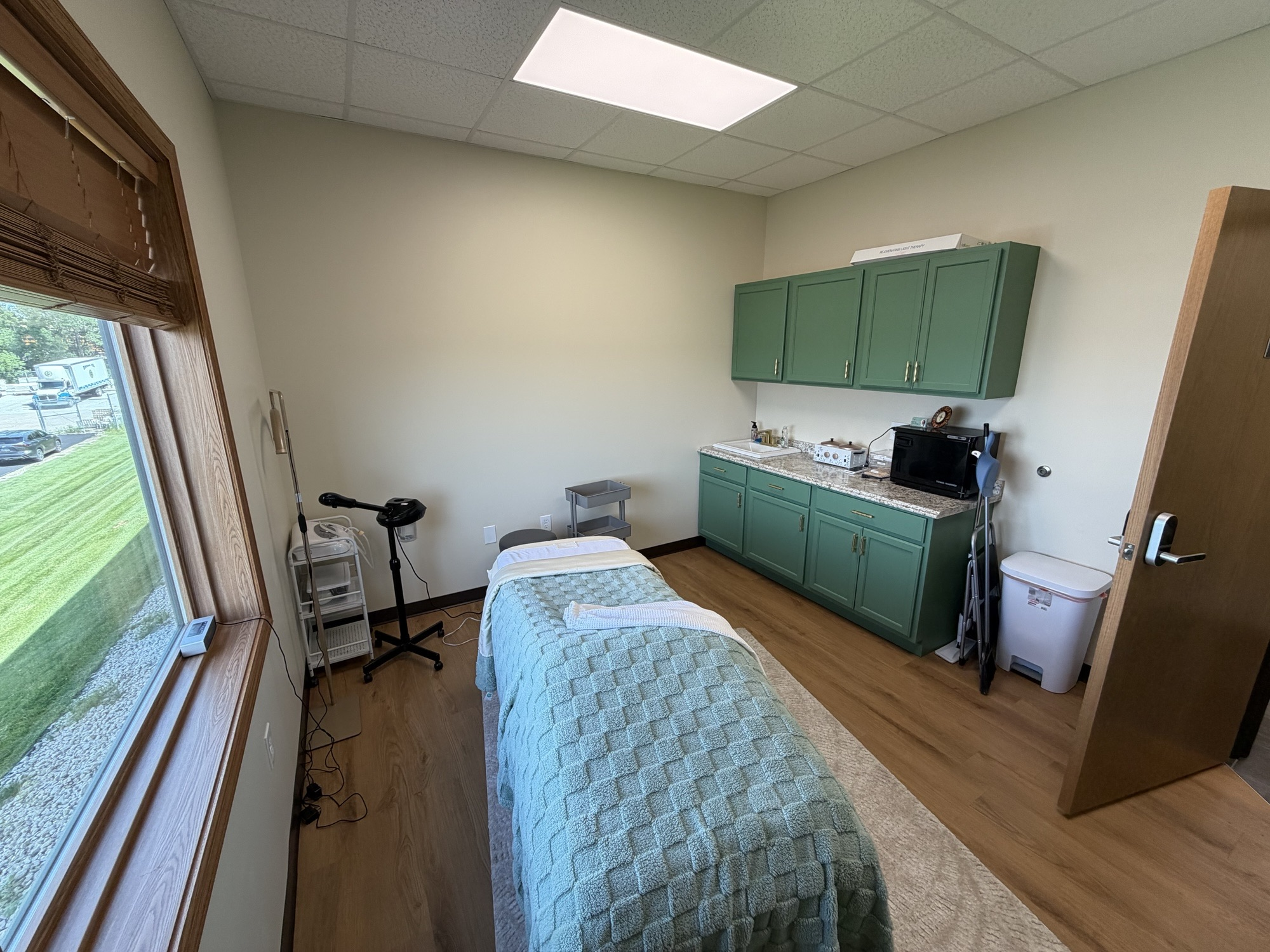 Suite 350 — wide view of the furnished treatment room with green built-in cabinets, sink, granite countertop on the right wall, massage table, and a large window overlooking grass and parking