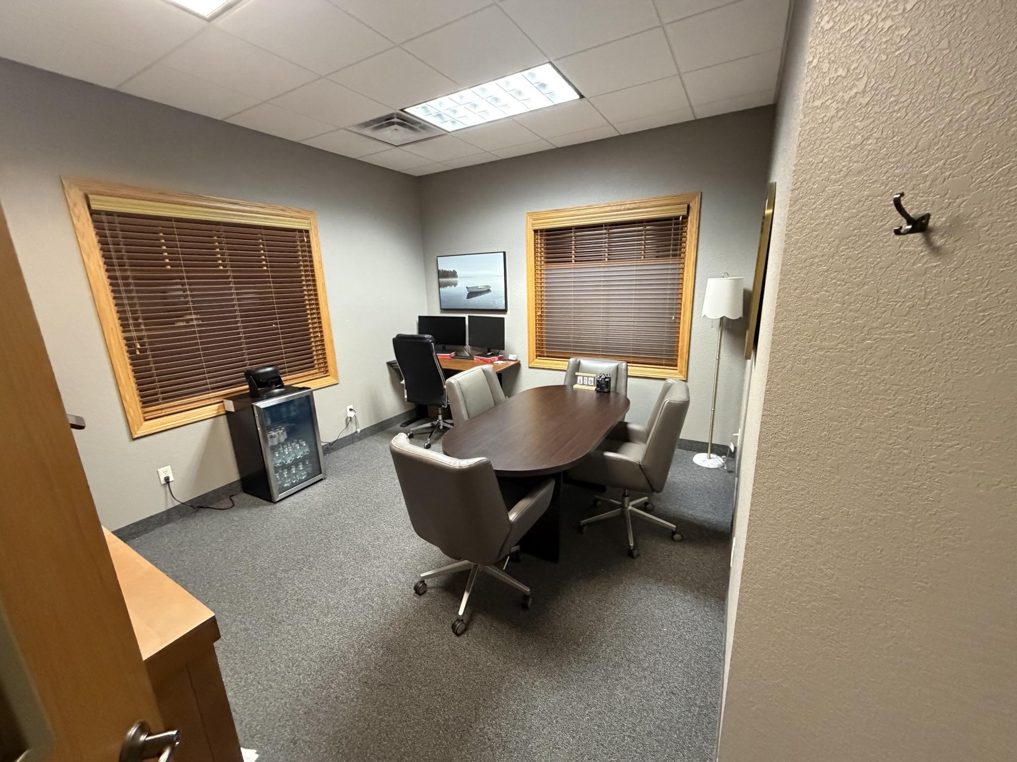 Suite 390 — corner executive office with two windows, oval meeting table with leather chairs, wall-mounted display, and floor lamp