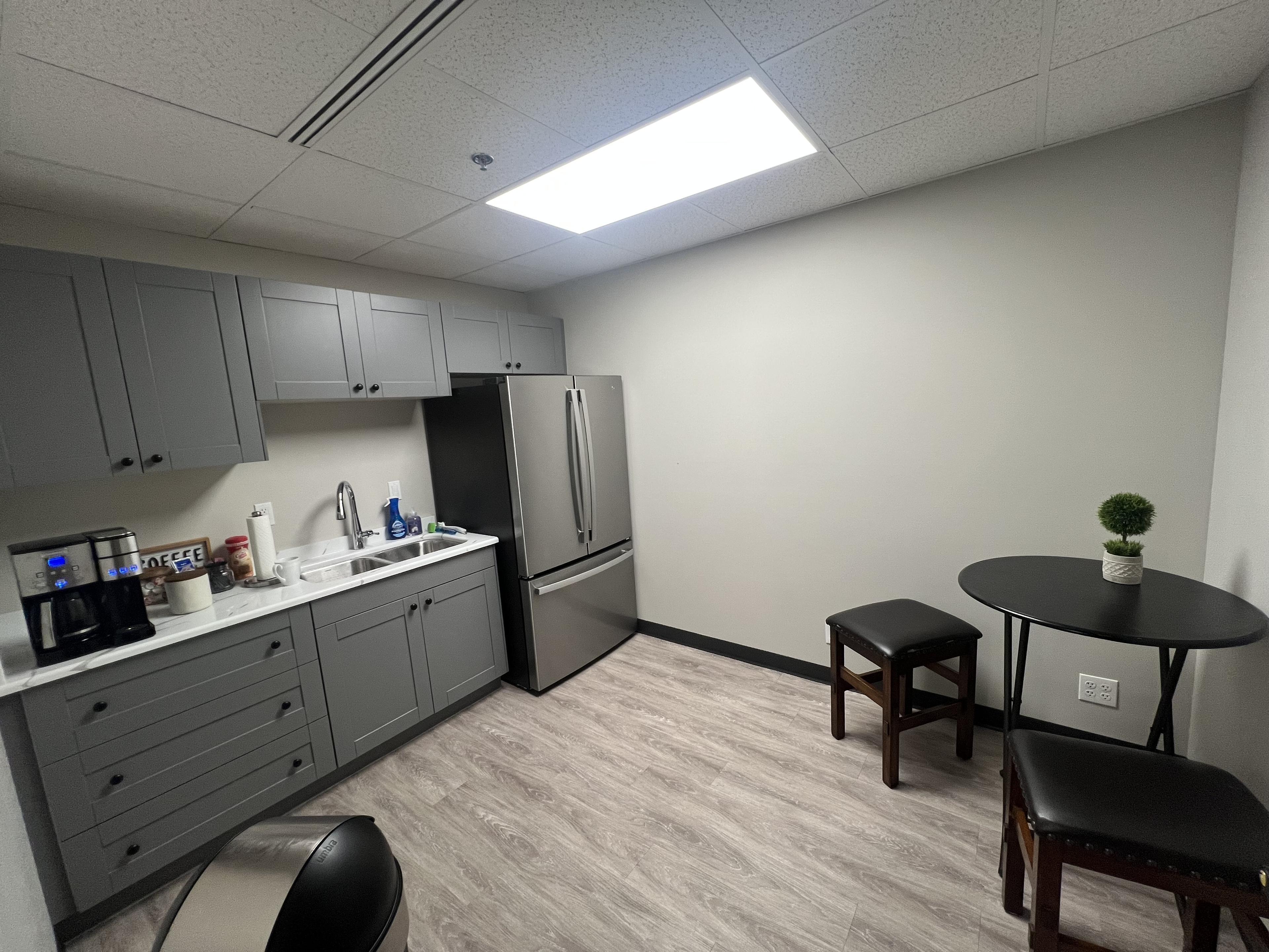 Modern break-room kitchen with gray shaker cabinets, stainless steel French-door fridge, sink, and stocked coffee bar