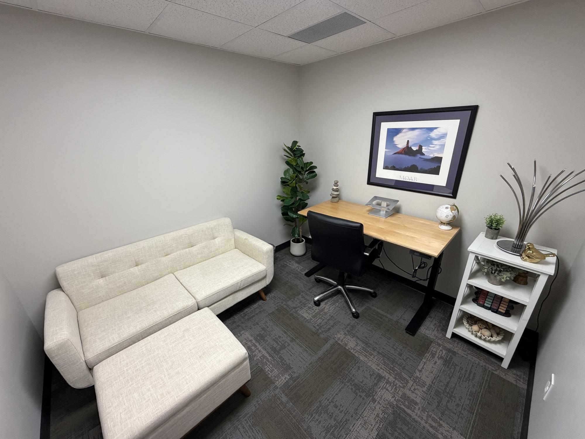 Office #1 at The Hub on 3rd — hourly private office with sit/stand desk, ergonomic chair, and a cream sofa seating area for client meetings